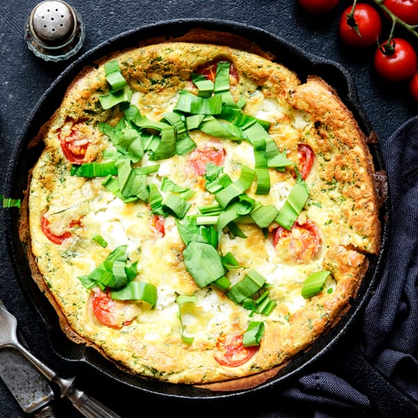 A frittata with tomatoes, cheese, and green herbs in a cast iron skillet.
