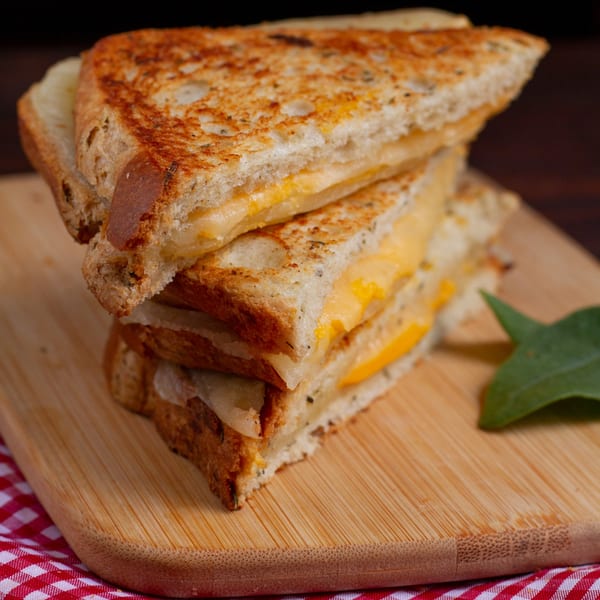Stack of three grilled cheese sandwiches with melted cheese on a wooden board with a red and white checkered napkin.