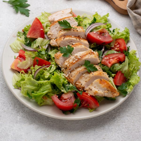 Grilled chicken breast slices with sesame seeds, fresh lettuce, red onion, and tomato on a white plate.